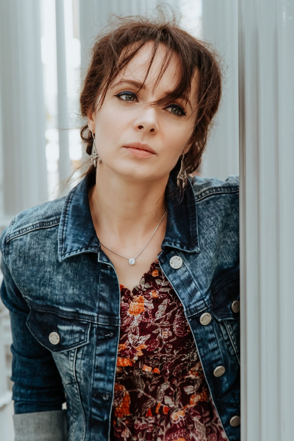 Olya Aman in a denim jacket and floral dress, leaning in a doorway