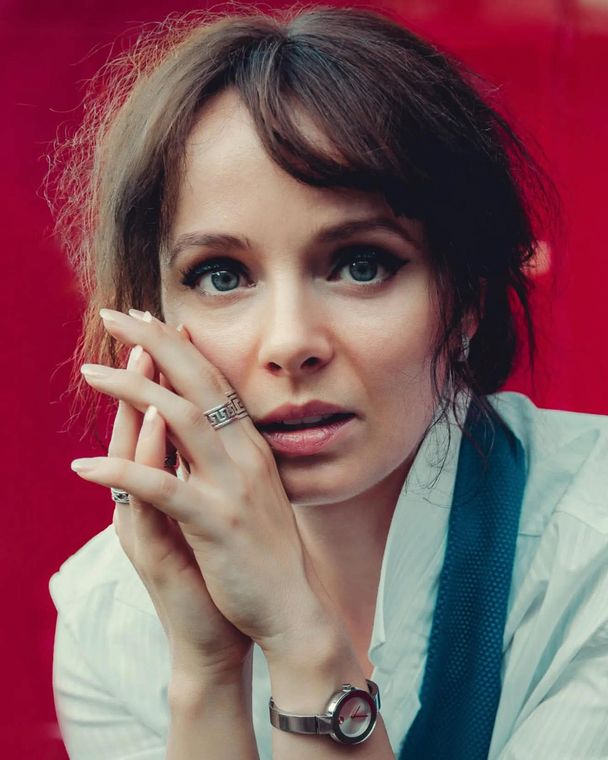 Olya against a red wall, fingertips at her cheek