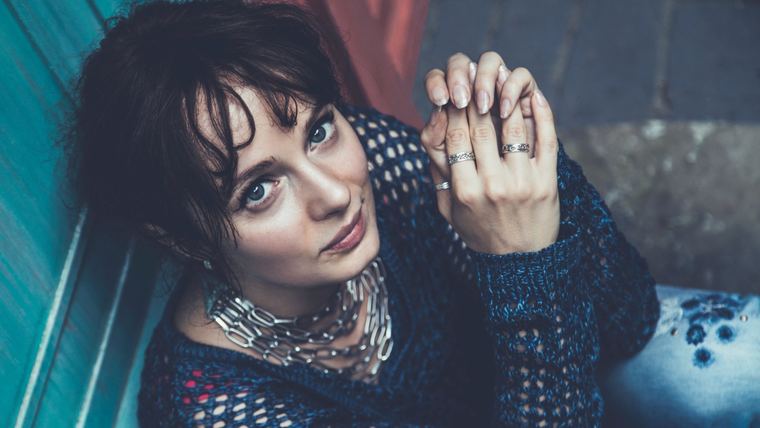 Olya in a dark knit sweater against teal, rings on her fingers